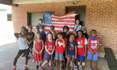 Summer program demand outpaces resources: Sixteen primary-schhol age children stand smiling in front of American flag held by two masked adult females in front of red brick wall