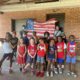 Summer program demand outpaces resources: Sixteen primary-schhol age children stand smiling in front of American flag held by two masked adult females in front of red brick wall