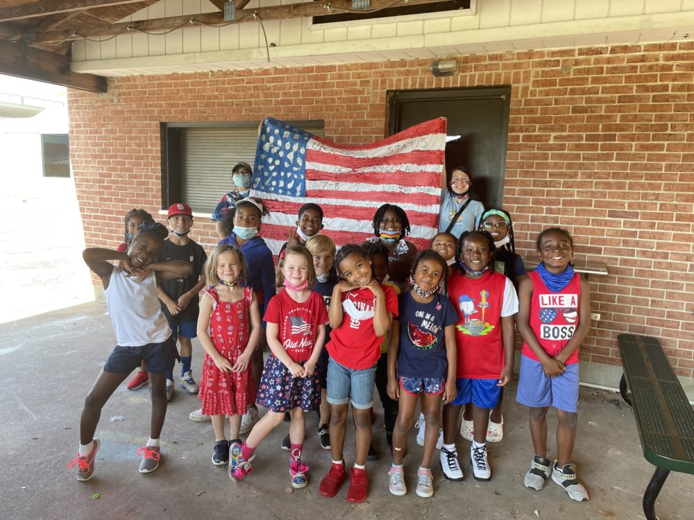 Summer program demand outpaces resources: Sixteen primary-schhol age children stand smiling in front of American flag held by two masked adult females in front of red brick wall