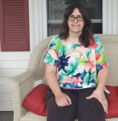 Disabled Roommates: Smiling woman with dark brown hair and dark-framed glasses w earing bright, floral blouse sits on wicker chair in front of white framed window with red shutters