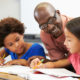 addressing student absenteeism, tutoring and afterschool programs: three pre-teen students sit working in notebooks while bald black man with dark glasses and red shirt explains lesson