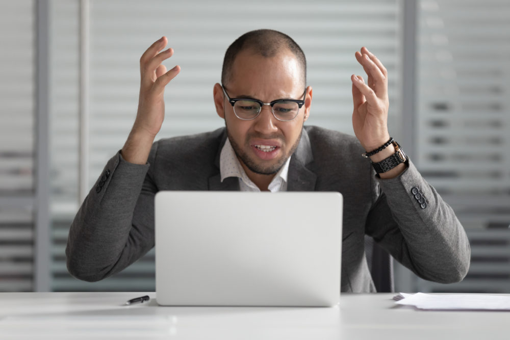 Grant makers hate 4 things: Black man with dark-rimmed glasses wearing gray suit and white shirt sits at silver laptop throwing hands in air with frustrated look