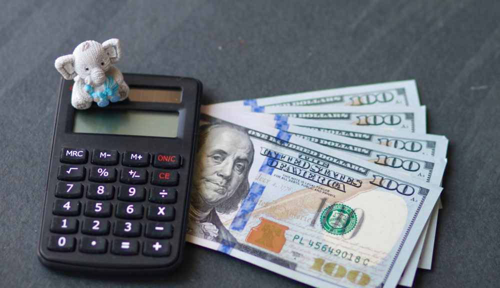 Child welfare solution: Calculator, five U.S. $100. bills and small stuffed elephant on dark gray tabletop