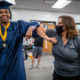 disabled people legal guardianship: Black woman with short braids in blue graduation robe and cap bumps elbows with woman wearing mask and grey t-shirt