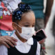 delta variant afterschool: young Elementary-age Black girl with black hair in top bun with large navy bow wearing dark navy and white top and face mask
