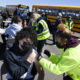 student vaccines: Several masked students sit at tables near yellow school buses with bright yellow jacketed health care workers administer vaccines