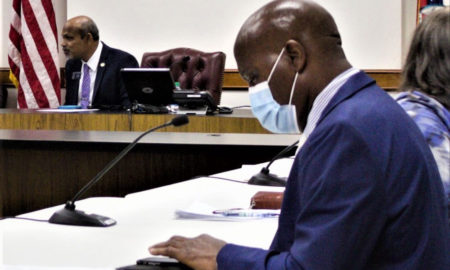 Outdoor Learning Study Committee: Bald black man in face mask wearing and blue suit sits at table in front of microphone with another black man in suit in the background.