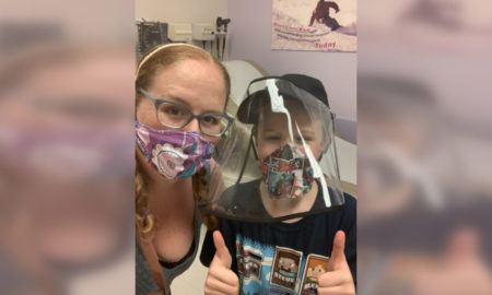 Disabled Speak Out: young boy in dark baseball cap and black t-sirt with plastic face shield and mask sits with blonde woman with face lanendar floral mask on hospital bed with medical gear in background.