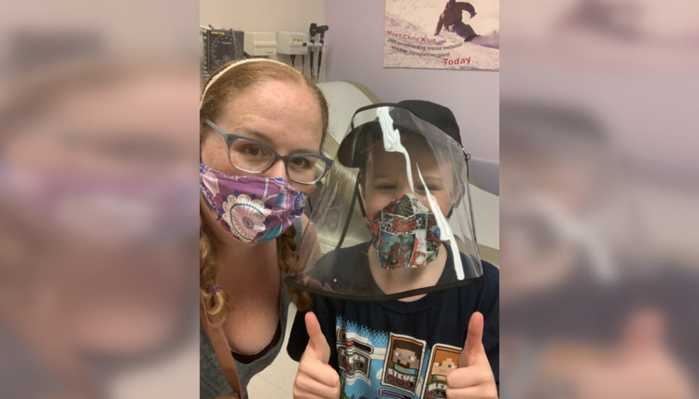 Disabled Speak Out: young boy in dark baseball cap and black t-sirt with plastic face shield and mask sits with blonde woman with face lanendar floral mask on hosital bed with medical gear in background.
