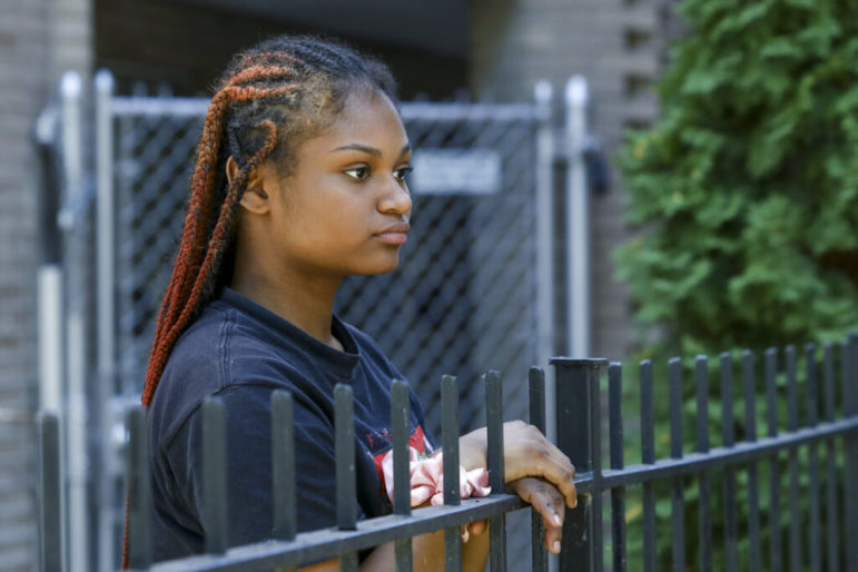 Police use excessive force with children: Black teen girl wearing black t-shirt with long red braids stands staring into the distance over top of black metal fence