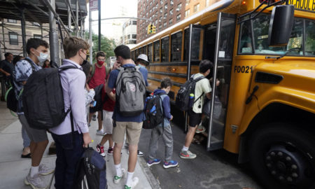 Afterschool Staff Shortage: Yellow school bus parked on street surrounded by tall buildings with several masked students wearing backpacks boarding bus.
