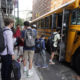 Afterschool Staff Shortage: Yellow school bus parked on street surrounded by tall buildings with several masked students wearing backpacks boarding bus.