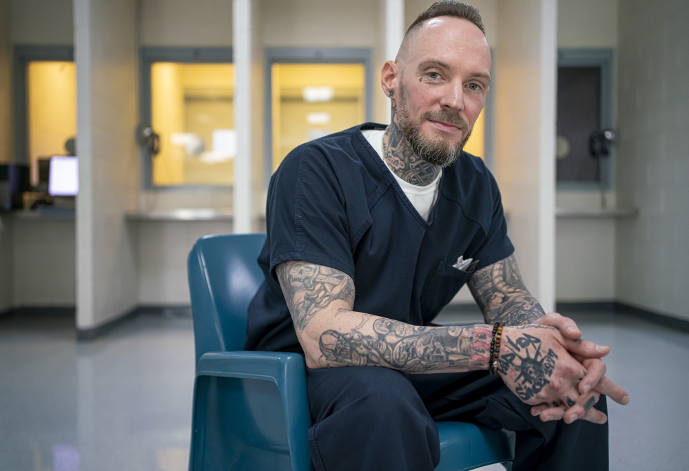 New Mexico jail: Michael Brown portrait with short light brown hair and beard smiles into camera sitting in turquoise plastic chair in prisoner visiting room wearing navy blue prison uniform with heavily black tatooed arms on lap and fingers interlaced.