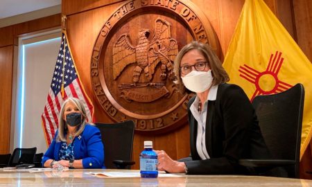 New Mexico supports tribal adoption: 2 women wearing masks and business clothessit in front of a wood wall with large, round, carved wood New Mexico State Seal with United States and New Mexico state flags to each side.