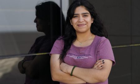 Texas abortion law shutting down courts for teens: dark haired, smiling woman wearing lavender t-shirt stands with arms crossed reflected in window behind her