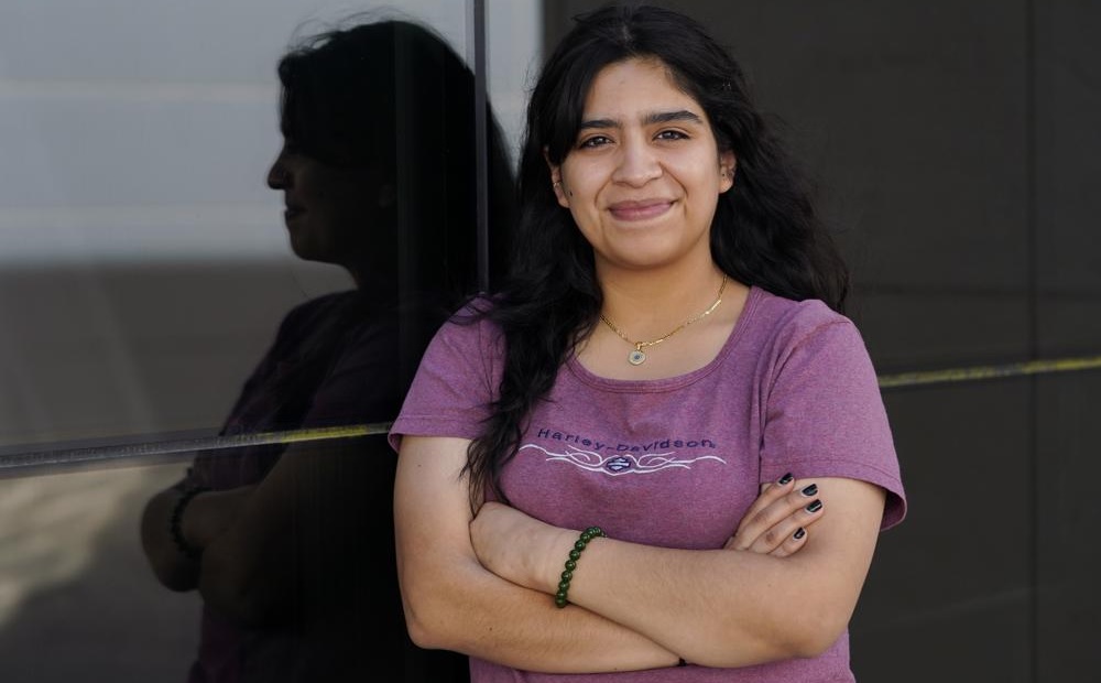 Texas abortion law shutting down courts for teens: dark haired, smiling woman wearing lavender t-shirt stands with arms crossed reflected in window behind her