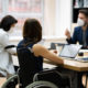 Disabled workers workplace: Three people sit around table with laptops - one person in a wheelchair - wearing face mask