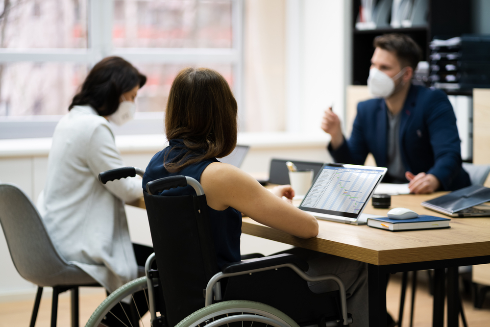 Disabled workers workplace: Three people sit around table with laptops - one person in a wheelchair - wearing face mask