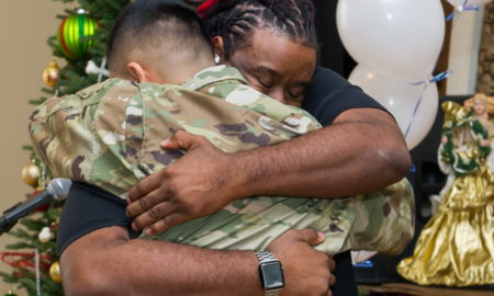 YMCA Urban Warriors: Black man with braids facing camera hugging man with short brown hair wearing camo military fatigues standing in front of a decorated Christmas tree