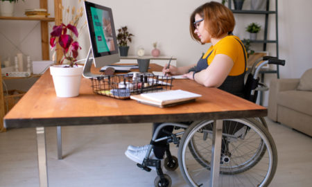Disabled remote workers: Young woman with short red hair and black frame glasses sits at desk in wheelchair with computer doing paperwork.