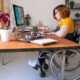 Disabled remote workers: Young woman with short red hair and black frame glasses sits at desk in wheelchair with computer doing paperwork.