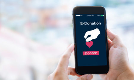 digital nonprofit fundraising: hand holding up black cell phone with red heart giving icon onscreen against blurry pastel background