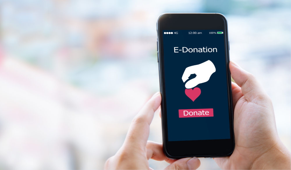 digital nonprofit fundraising: hand holding up black cell phone with red heart giving icon onscreen against blurry pastel background
