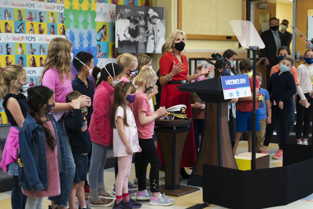 COVID-19 school vaccinations: Blonde woman in red dress and black mask stands speaking at wood podium with blue and white "Vaccines.gov" sign in front or podium, surrounded by young children wearing masks.