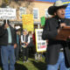 NM k-12 Curriculum: Man in cowboy hat,blue jeans and brown jacket speaks to crowd gathered outdoors holding protest signs.