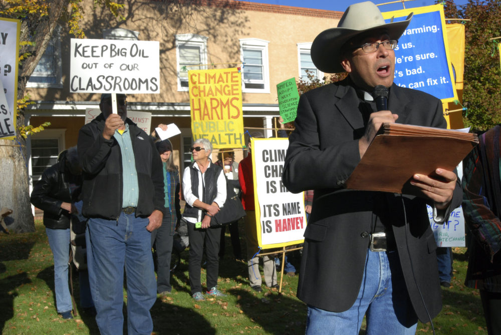 NM k-12 Curriculum: Man in cowboy hat,blue jeans and brown jacket speaks to crowd gathered outdoors holding protest signs.