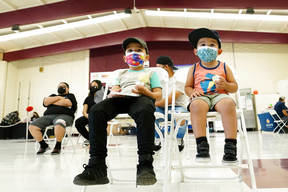 Youth vaccine disparities: Two young boys and several parents all wearing masks, sit on folding chairs scattered about inside a large white and red school auditorium