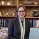 New Mexico child welfare department: Barbara Vigil brown -haired woman with dark glasses in navy jacket with white scarf smiles into the camera standing in front of light wood book shelves