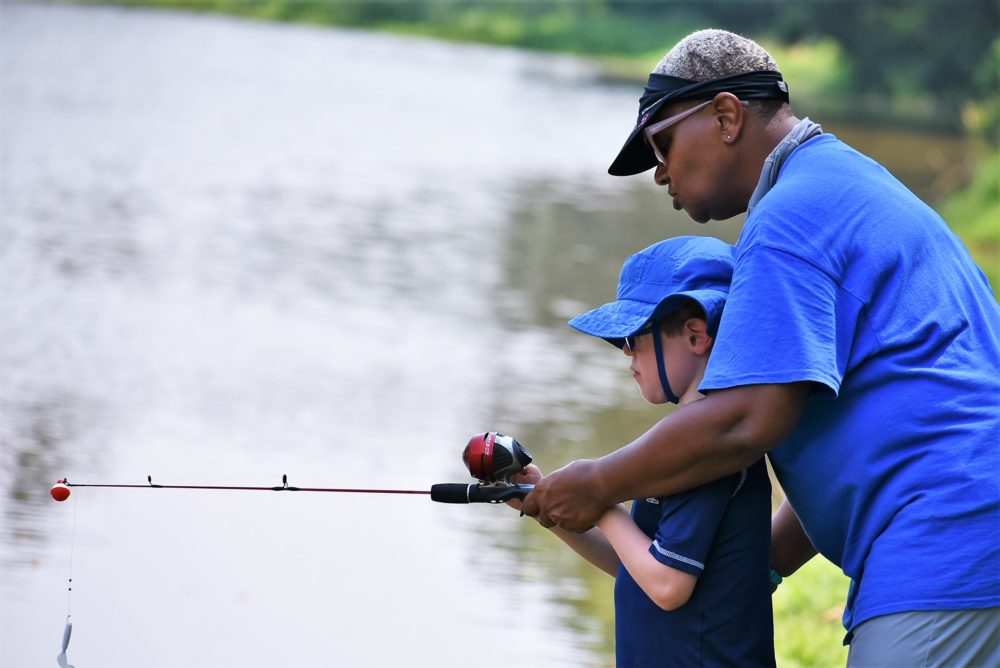 summer camp: Black man in baseball cap teaching young child how to hold fishing pole