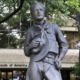 Boy Scouts Camps Sell-off: Gray metal statue of teen boy in Scout uniform stands in front of leafy green trees