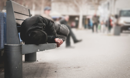 NY Homeless Payments: Person in dark clothes sits cross-legged on city bench huddled over