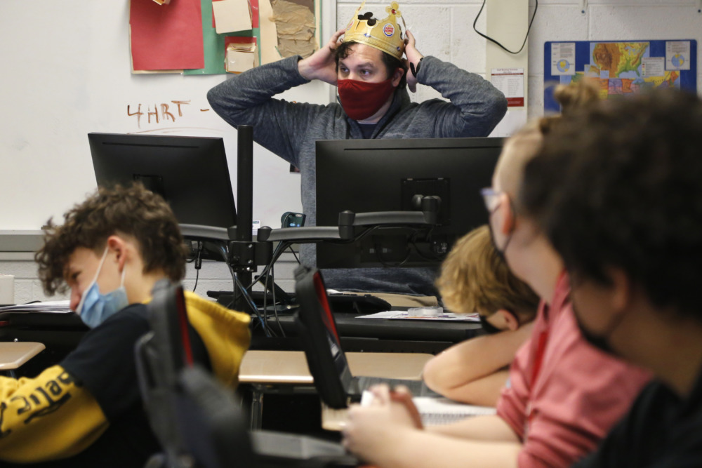 School therapy program: Man with dark hair wearing yellow paper crown, red mask and grey sweater sits i at desk with computer monitors in front of four pre-teen students wearing masks sitting at their desks.