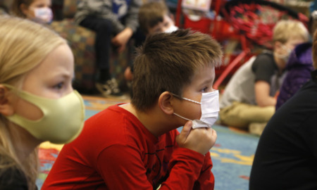 School therapy program: two elementary students wearing masks sit on floor with many other students sitting cross-legged in the background