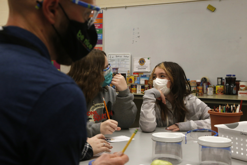 Schol therapy program: Two young girls with long brown hair wearing masks sit at table with adult wearing mask and glasses.