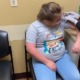 Down Syndrome vaccine effects: Young girl with blonde hair wearing glasses, light blue pants and light blue t-shirt, sits in a chair while someone wearing gloves and a dark clothes gives her a shot in upper left arm.