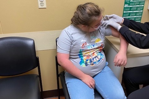 Down Syndrome vaccine effects: Young girl with blonde hair wearing glasses, light blue pants and light blue t-shirt, sits in a chair while someone wearing gloves and a dark clothes gives her a shot in upper left arm.