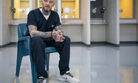 Sentencing reform: Michael Brown, brown-haired man with heavily tattooed arms in navy prison uniform sits with arms on knees and clasped hands in room with telephone cubbies along back wall