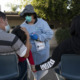 COVID Kids Hospitalization Surge: 2 adults with backs to camera sit holding young child while masked adult in medical gown weaing hat and sunglasses takes nasal swab of child in outdoor parking lot.