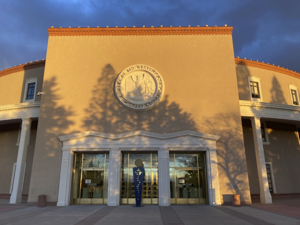 New Mexico child welfare: New Mexico beige state building entrance with white state seal and tree shadows