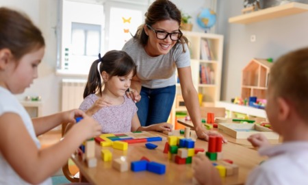 early care and education: female teacher with glasses leans over table helping children play with blocks
