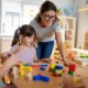 early care and education: female teacher with glasses leans over table helping children play with blocks