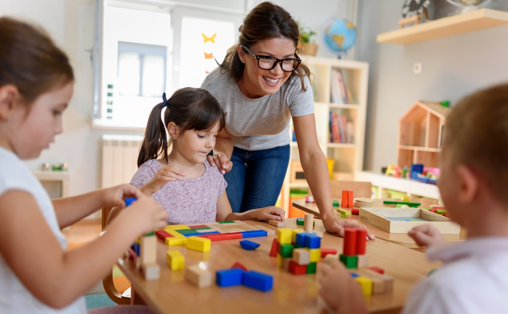 early childhood care and education: female teacher with glasses leans over table helping children play with blocks