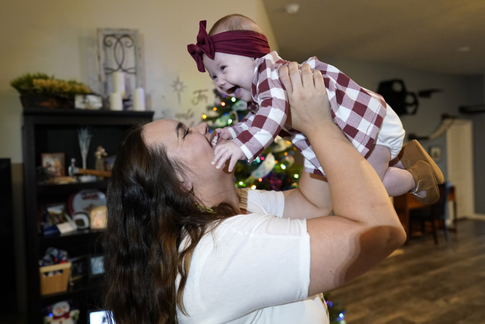 Abortion and Adoption: Young, dark-haired woman in white top holds baby aloft in the air. Baby wears a red plaid dress and red bow headband