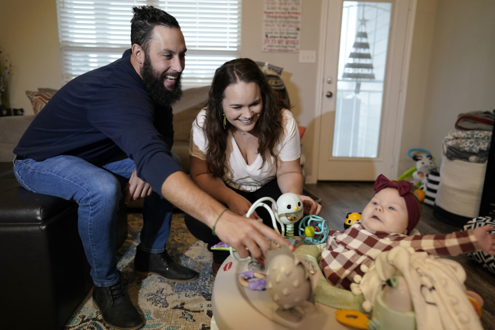 Abortion and Adoption: Two dark-haired male and female adults sit on couch leaning over and smiling at baby in red plaid dress in baby carrier on floor.