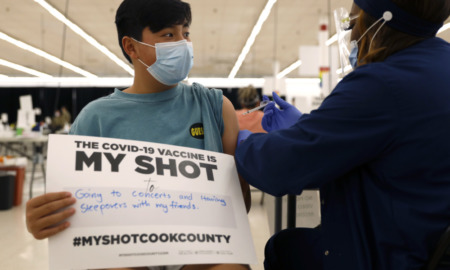Omicron Booster: Dark-haired young teen wearing mask holds sign "My Shot" as he receives COVID vaccine from adult wearing mask and long-sleeved navy uniform.