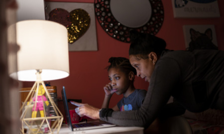 Digital divide virus: Older Black woman leans over shoulder of young black girl sitting at desk looking at laptop screen